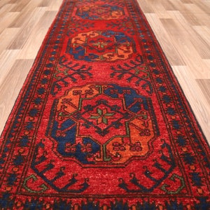 May include: A red, blue, and green patterned rug with a traditional design. The rug has a dark blue border and is laid out on a wooden floor.