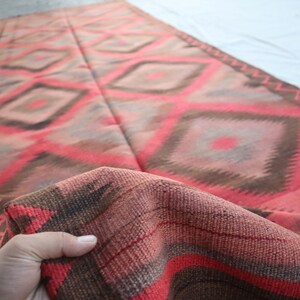 May include: A red, brown, and gray woven rug with a geometric diamond pattern. The rug is folded in half and the edges are fringed.