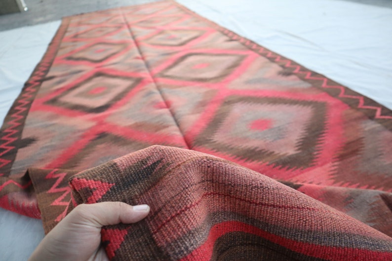May include: A red, brown, and gray woven rug with a geometric diamond pattern. The rug is folded in half and the edges are fringed.