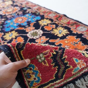 May include: A detailed shot of a Persian rug with a dark blue background and a vibrant floral pattern in orange, yellow, and blue. The rug's red border and fringed edge are visible, showcasing its intricate design.