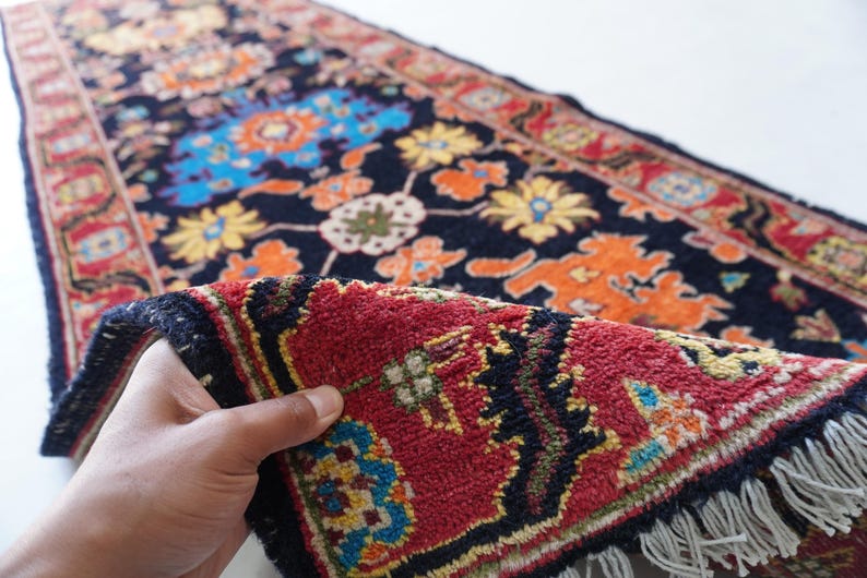 May include: A detailed shot of a Persian rug with a dark blue background and a vibrant floral pattern in orange, yellow, and blue. The rug's red border and fringed edge are visible, showcasing its intricate design.