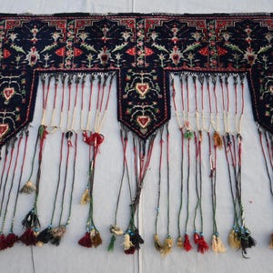 Puede incluir: Textil rectangular azul oscuro con intrincados patrones florales y geom&eacute;tricos en rojo, blanco y verde. El textil tiene una serie de borlas colgantes en varios colores, incluyendo rojo, negro y crema. El dise&ntilde;o presenta motivos en forma de coraz&oacute;n.