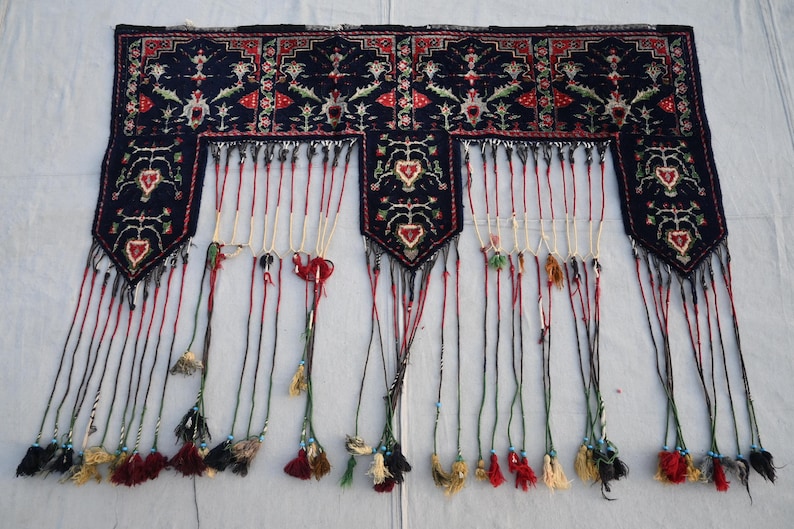 Puede incluir: Textil rectangular azul oscuro con intrincados patrones florales y geom&eacute;tricos en rojo, blanco y verde. El textil tiene una serie de borlas colgantes en varios colores, incluyendo rojo, negro y crema. El dise&ntilde;o presenta motivos en forma de coraz&oacute;n.