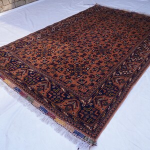 May include: A rectangular, hand-woven rug with an intricate pattern. The rug features a predominantly rust-colored field with a detailed floral design. Dark blue borders frame the central pattern, and the rug has fringed edges.