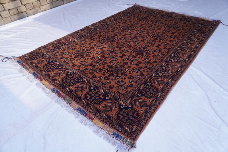 May include: A rectangular, hand-woven rug with an intricate pattern. The rug features a predominantly rust-colored field with a detailed floral design. Dark blue borders frame the central pattern, and the rug has fringed edges.