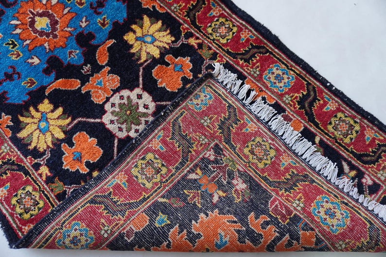 May include: An intricately patterned rug with a dark blue background and a vibrant floral design in shades of orange, yellow, and blue. The rug's border is red with a repeating geometric pattern, and it has a fringed edge.