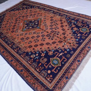 May include: An antique Persian rug with a central diamond design in a field of rust-orange. The rug features a navy blue border with intricate floral and geometric patterns. The rug has a fringed edge and is displayed on a white surface.