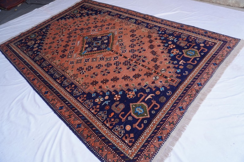 May include: An antique Persian rug with a central diamond design in a field of rust-orange. The rug features a navy blue border with intricate floral and geometric patterns. The rug has a fringed edge and is displayed on a white surface.
