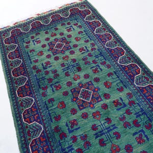 May include: A rectangular, hand-woven rug with a teal background and intricate patterns. The rug features a central design with red and blue geometric shapes, surrounded by a detailed border. The rug's dimensions are not visible.