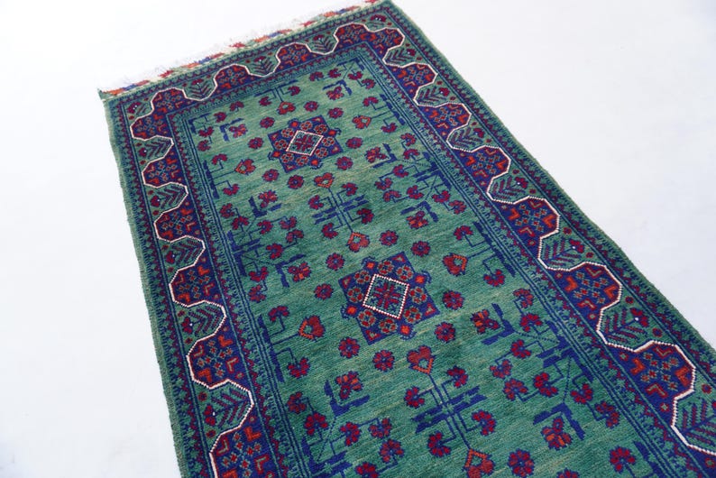 May include: A rectangular, hand-woven rug with a teal background and intricate patterns. The rug features a central design with red and blue geometric shapes, surrounded by a detailed border. The rug's dimensions are not visible.