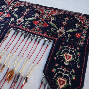 Puede incluir: Primer plano de una alfombra azul oscuro con intrincados dise&ntilde;os florales en rojo, verde y blanco. La alfombra tiene un borde con flecos con hilos rojos y crema, y borlas decorativas.