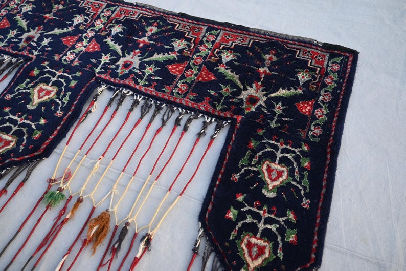 Puede incluir: Primer plano de una alfombra azul oscuro con intrincados dise&ntilde;os florales en rojo, verde y blanco. La alfombra tiene un borde con flecos con hilos rojos y crema, y borlas decorativas.