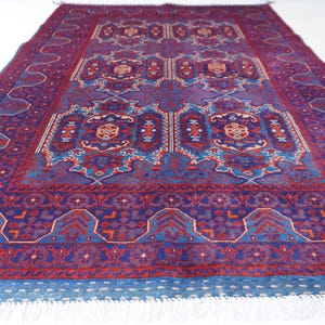May include: A large, rectangular rug with an intricate, symmetrical design. The rug features a deep burgundy field with blue and red geometric patterns. A blue border with white fringe completes the design. The rug is suitable for a living room or bedroom.