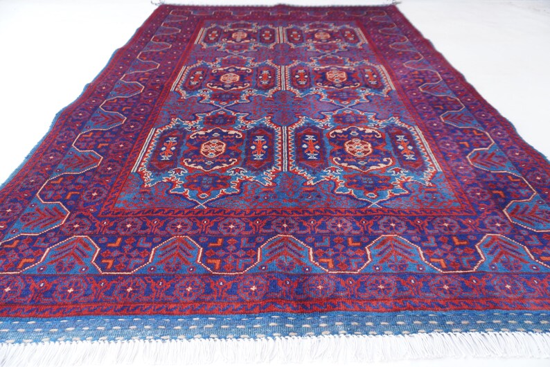 May include: A large, rectangular rug with an intricate, symmetrical design. The rug features a deep burgundy field with blue and red geometric patterns. A blue border with white fringe completes the design. The rug is suitable for a living room or bedroom.