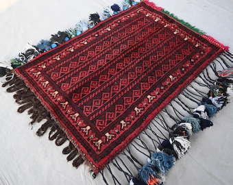 May include: A rectangular, hand-woven rug with a dark red and black geometric pattern. The rug has a fringe of tassels in various colors, including blue, black, and white. The rug is likely used as a decorative item.
