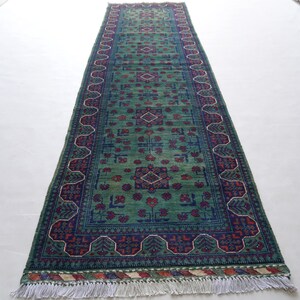 May include: A rectangular green rug with a central pattern of red and blue floral designs. The rug has a dark blue border with a repeating pattern and white fringe on the ends. The rug is displayed on a white surface.