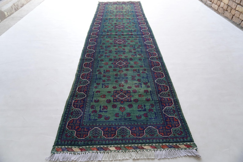 May include: A rectangular green rug with a central pattern of red and blue floral designs. The rug has a dark blue border with a repeating pattern and white fringe on the ends. The rug is displayed on a white surface.