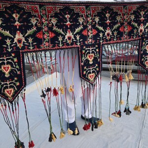 K&ouml;nnte beinhalten: Dunkelblaues Textil mit detailliertem Blumenmuster in Rot, Gr&uuml;n und Wei&szlig;. Das Textil hat einen Fransenrand mit Quasten in verschiedenen Farben. Das Design umfasst herzf&ouml;rmige Motive und komplizierte florale Elemente.