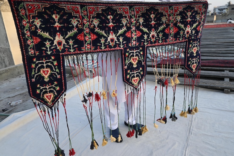 K&ouml;nnte beinhalten: Dunkelblaues Textil mit detailliertem Blumenmuster in Rot, Gr&uuml;n und Wei&szlig;. Das Textil hat einen Fransenrand mit Quasten in verschiedenen Farben. Das Design umfasst herzf&ouml;rmige Motive und komplizierte florale Elemente.
