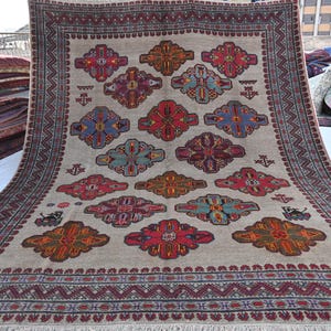 May include: A large, rectangular rug with a beige background and a repeating pattern of colorful floral motifs. The border is a deep red with geometric designs. The rug has a fringed edge and is likely made of wool.