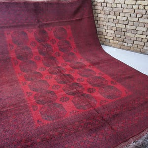 May include: A burgundy-colored rug with a central red panel featuring a repeating pattern of dark shapes. The rug has a detailed border and fringe along one edge. The rug is displayed on a white surface.