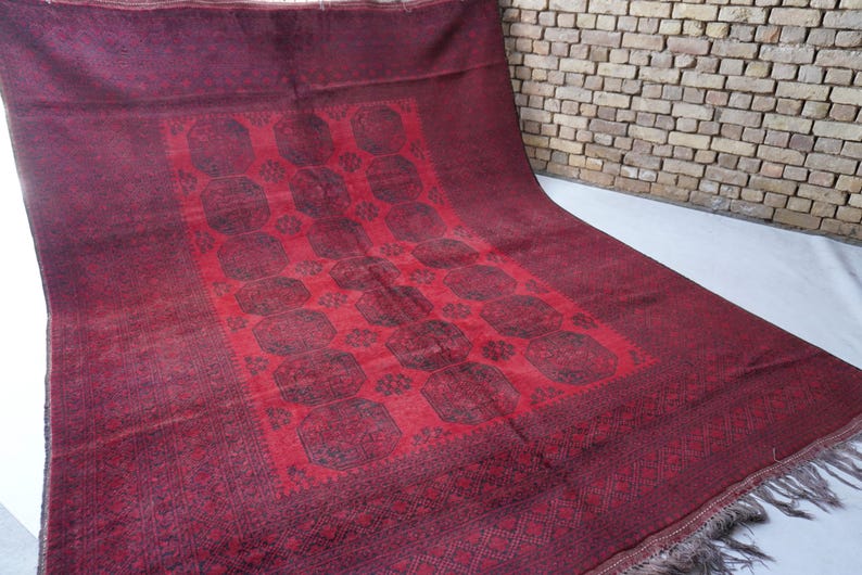 May include: A burgundy-colored rug with a central red panel featuring a repeating pattern of dark shapes. The rug has a detailed border and fringe along one edge. The rug is displayed on a white surface.