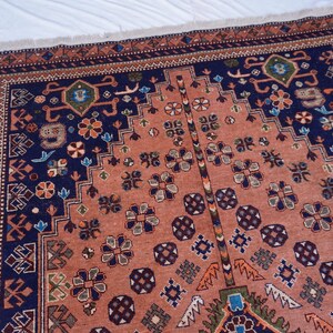 May include: A detailed, rectangular rug with a central peach-colored field and a navy blue border. The rug features intricate floral and geometric patterns in shades of blue, green, and brown. The edges have a fringed trim.