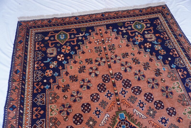 May include: A detailed, rectangular rug with a central peach-colored field and a navy blue border. The rug features intricate floral and geometric patterns in shades of blue, green, and brown. The edges have a fringed trim.