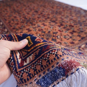 May include: A detailed, hand-knotted rug with a warm, earthy color palette. The rug features intricate patterns in shades of orange, blue, and red. The fringe is white. The rug is being held up to show its texture and detail.