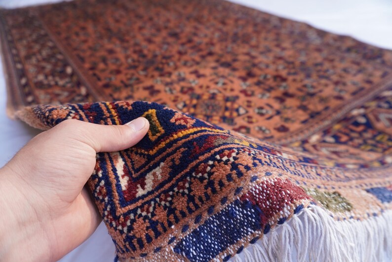 May include: A detailed, hand-knotted rug with a warm, earthy color palette. The rug features intricate patterns in shades of orange, blue, and red. The fringe is white. The rug is being held up to show its texture and detail.
