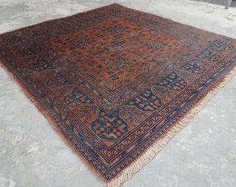 May include: A square area rug featuring a rust-orange field with a dark blue geometric border. The rug displays a repeating pattern of intricate floral and medallion motifs in dark blue and black, finished with short beige fringe on two sides.