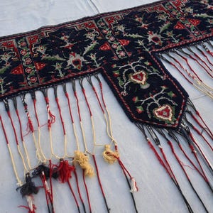 K&ouml;nnte beinhalten: Ein dunkelblaues Textil mit aufwendigen Blumenmustern in Rot, Gr&uuml;n und Wei&szlig;. Die R&auml;nder sind mit langen, farbenfrohen Quasten verziert. Dieses dekorative St&uuml;ck k&ouml;nnte ein Wandbehang oder ein Textilakzent sein.