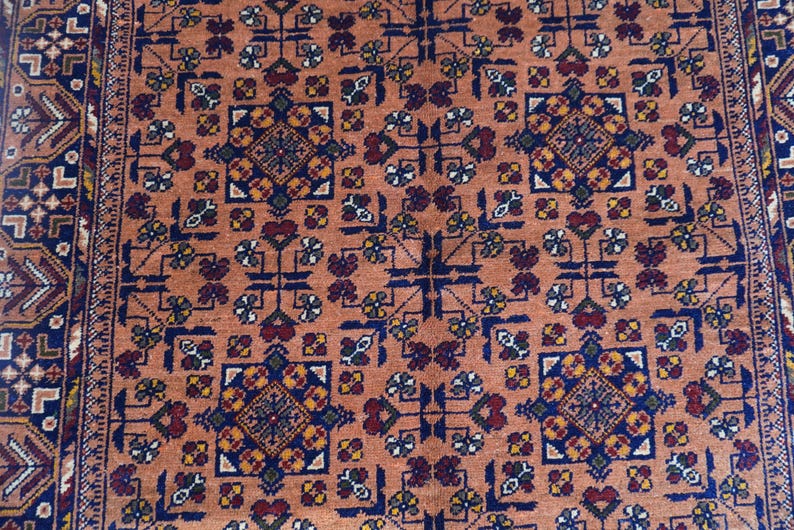 May include: A detailed close-up of a rug with a repeating geometric pattern. The rug features a warm, peach-colored background with a complex design in navy blue, burgundy, and gold. The border is a darker blue with intricate detailing.
