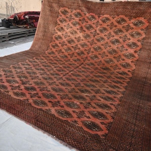 Könnte beinhalten: Ein großer, rechteckiger Teppich mit einem sich wiederholenden Rautenmuster in Rost- und Brauntönen. Der Teppich hat einen dunkelbraunen Rand mit geometrischen Mustern. Der Teppich besteht aus einem gewebten Material und scheint handgefertigt zu sein.