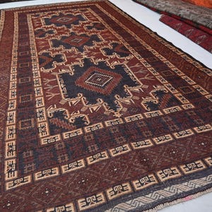 May include: A rectangular, hand-woven rug with a geometric pattern in shades of burgundy, navy, and beige. The rug features a central diamond design and a border with repeating motifs. The rug is likely made of wool and is suitable for a living room or bedroom.