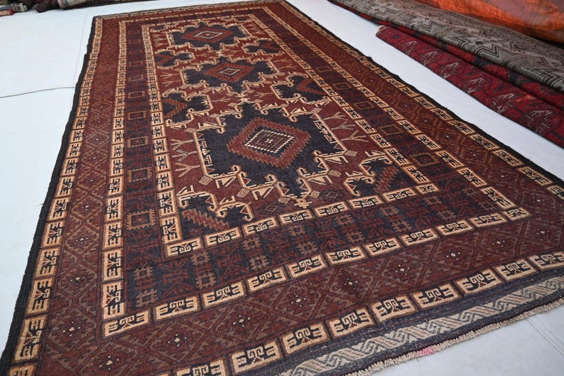 May include: A rectangular, hand-woven rug with a geometric pattern in shades of burgundy, navy, and beige. The rug features a central diamond design and a border with repeating motifs. The rug is likely made of wool and is suitable for a living room or bedroom.