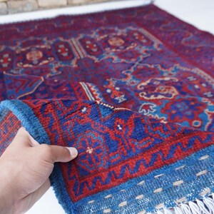 May include: A hand-woven rug with a rich, intricate design. The rug features a deep red and blue color palette with detailed patterns. The border is a vibrant blue with a fringe. The rug is folded, revealing its texture and craftsmanship.