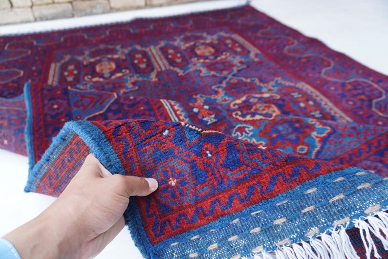 May include: A hand-woven rug with a rich, intricate design. The rug features a deep red and blue color palette with detailed patterns. The border is a vibrant blue with a fringe. The rug is folded, revealing its texture and craftsmanship.