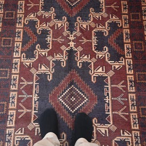 May include: A rectangular rug with a geometric pattern in shades of burgundy, navy blue, and beige. The rug features a central diamond design and repeating border motifs. The rug is viewed from above, with two feet visible.
