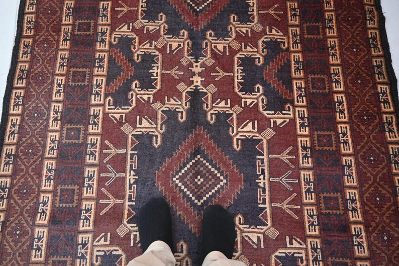 May include: A rectangular rug with a geometric pattern in shades of burgundy, navy blue, and beige. The rug features a central diamond design and repeating border motifs. The rug is viewed from above, with two feet visible.