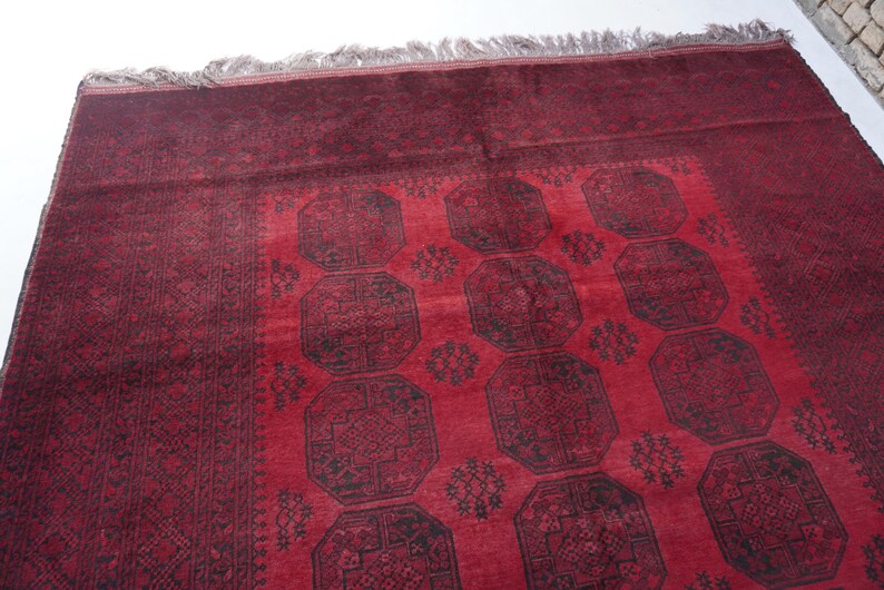 May include: A red and black patterned rug with a central design of repeating octagonal shapes. The rug has a dark red border and a lighter red central field. The fringe is a light gray color.
