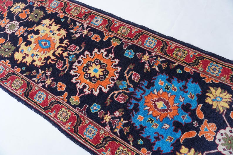 May include: A detailed shot of a dark blue runner rug with a vibrant, intricate floral pattern. The design features orange, yellow, and blue floral motifs, framed by a red border. The rug's design is symmetrical and ornate, suitable for home decor.