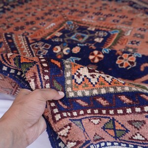 May include: A detailed shot of a folded, patterned rug. The rug features a complex design with a navy blue border and a peach-colored field. Intricate floral and geometric patterns in orange, green, and white adorn the rug's surface. The rug is held up, showcasing its texture.