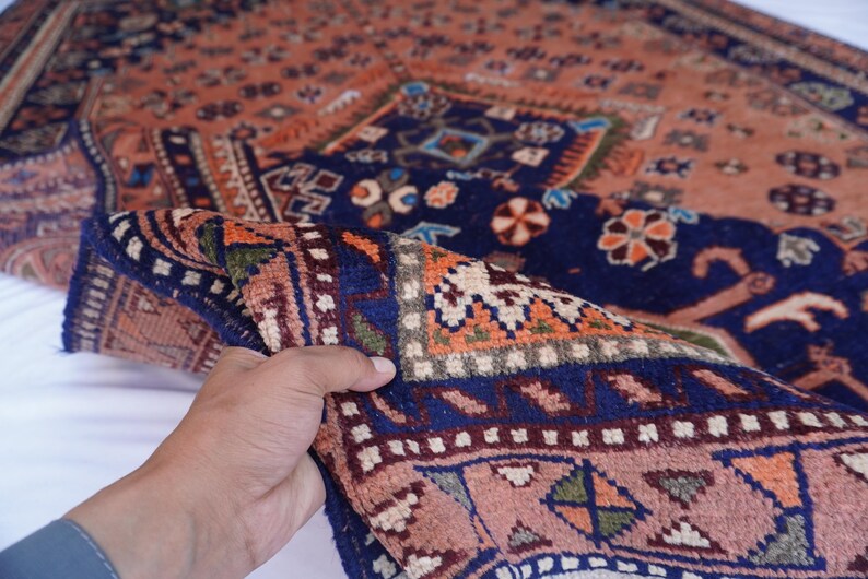 May include: A detailed shot of a folded, patterned rug. The rug features a complex design with a navy blue border and a peach-colored field. Intricate floral and geometric patterns in orange, green, and white adorn the rug's surface. The rug is held up, showcasing its texture.