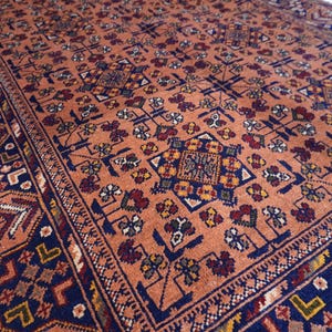 May include: An antique rug with a detailed, repeating pattern. The rug features a warm, peach-colored background with intricate designs in navy blue, red, yellow, and white. The border has a geometric pattern. This rug is suitable for home decor.