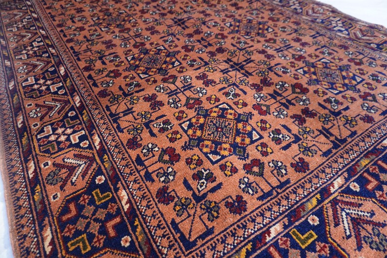 May include: An antique rug with a detailed, repeating pattern. The rug features a warm, peach-colored background with intricate designs in navy blue, red, yellow, and white. The border has a geometric pattern. This rug is suitable for home decor.