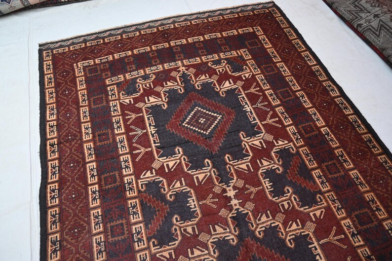 May include: A rectangular, hand-woven rug with a central geometric design in dark blue and burgundy. The rug features intricate patterns and borders in shades of beige, brown, and black. The rug's design is symmetrical and traditional.