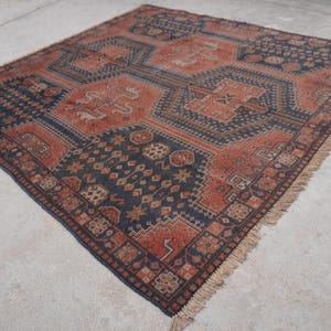 Pu&ograve; includere: Tappeto quadrato con un motivo geometrico nei toni del ruggine, blu navy e beige. Il tappeto presenta un disegno centrale con forme ripetute e un bordo sfrangiato. Il tappeto &egrave; posto su una superficie di cemento.