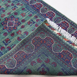 May include: A teal-colored rug with a detailed pattern of red and navy blue floral and geometric designs. The rug's edge is visible, revealing a fringed border and the underside with a similar pattern.