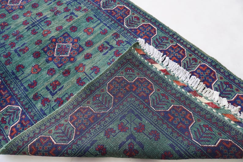 May include: A teal-colored rug with a detailed pattern of red and navy blue floral and geometric designs. The rug's edge is visible, revealing a fringed border and the underside with a similar pattern.